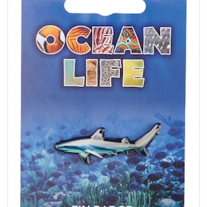 Könnte beinhalten: Eine silberfarbene Hai-Anstecknadel auf einer blauen Trägerkarte. Die Karte zeigt oben den Text OCEAN LIFE in großen, gemusterten Buchstaben und unten die Worte PIN BADGE vor einer Unterwasserszene mit Fischen und Korallen im Hintergrund.
