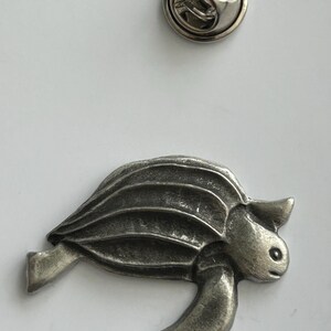 May include: A silver-coloured metal turtle pin with a textured shell and flippers. The pin has a detailed design and a classic pin back. A separate silver-coloured pin back is also visible.