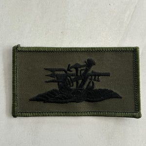 Puede incluir: Un parche rectangular de tela verde oliva con un emblema bordado en verde oscuro en el centro. El emblema presenta un diseño estilizado de un tridente, un martillo y una llave cruzados sobre una base de follaje, todo cosido con hilo verde.