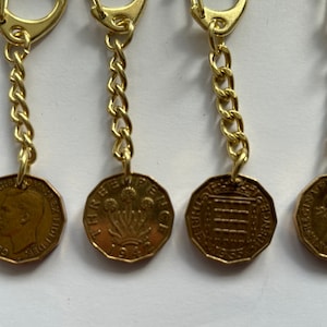 May include: Four gold keychains with different coin designs. Each keychain features a gold chain and a round coin with a different design. The coins feature a profile of a person, a floral design, a building, and a crown.