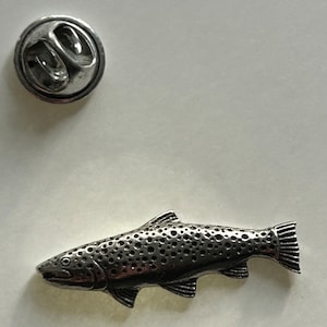Może przedstawiać: Srebrna metalowa spinka w kształcie pstrąga z detaliczną, fakturowaną powierzchnią. Ryba ma cętkowany wzór i lekko otwarte usta.