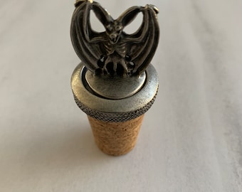 Puede incluir: Un tapón de botella de plata con un diseño de murciélago detallado. El murciélago tiene las alas extendidas y una expresión amenazante.