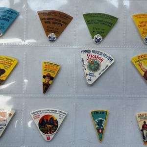 Peut inclure: Une collection d'étiquettes de fromage vintage dans un protège-feuilles en plastique transparent. Les étiquettes sont triangulaires et présentent divers motifs et textes, notamment "Swiss Process Cream Gruyere", "Finnish Process-Gruyere Dorsay" et "Du Chino".