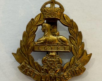 May include: A gold-colored metal cap badge with a crown, a sphinx, a wreath of leaves, and the text "EAST LANCASHIRE" and "EGYPT".