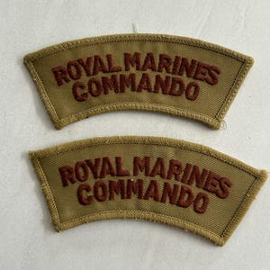 May include: Two tan fabric patches, each embroidered with "ROYAL MARINES COMMANDO" in brown. The patches are curved and likely represent military insignia or badges.
