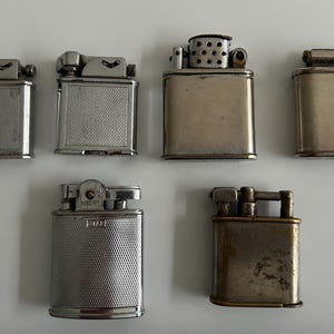 May include: Six vintage silver lighters, some with a textured finish, arranged on a white surface.
