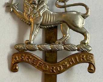 May include: A silver-colored metal emblem featuring a lion with a star-shaped object in its paw. The emblem has a banner below with the word "HEREFORDSHIRE" in gold lettering. The emblem is likely a military or heraldic badge.