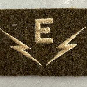May include: A rectangular olive green felt patch featuring an embroidered beige letter E centered above two symmetrical beige lightning bolt symbols that point downwards and outwards from the center of the patch in a V-shaped arrangement.