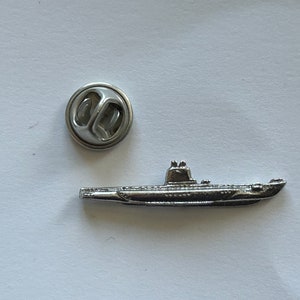 Puede incluir: Un pin de metal plateado de un submarino con un cierre de pin de metal plateado.