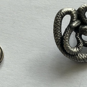 Puede incluir: Un broche de metal plateado con un diseño de serpiente enroscada. El broche tiene una textura detallada y un pasador con cierre. La cabeza de la serpiente es visible en el centro del enroscamiento. Es probable que sea un accesorio de moda.