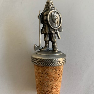 May include: A silver bottle stopper with a Viking warrior figure. The warrior holds an axe and shield, and is mounted on a cork base. The stopper is designed for standard wine bottles.