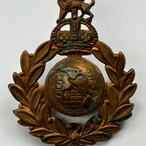May include: A vintage bronze-toned metal emblem featuring a globe encircled by a laurel wreath. A crown and lion sit atop the globe, with the continents of Europe, Asia, and Africa visible. The emblem has a weathered appearance.