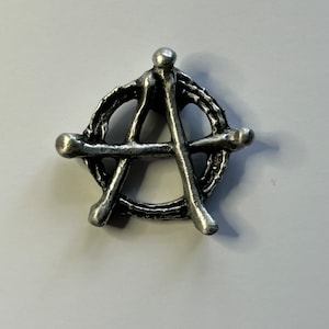May include: A silver-coloured metal pin in the shape of the anarchy symbol. The symbol is made of a circle with a stylized A inside.
