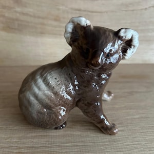 Op de afbeelding: Een bruin en wit keramiek koalabeeldje. De koala zit met zijn hoofd naar de zijkant gedraaid en zijn oren omhoog.