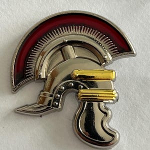 Könnte beinhalten: Eine römische Helmnadel aus Silber und Gold mit einem roten Kamm. Der Helm ist mit erhabenen Linien und Punkten verziert. Der Kamm ist eine gebogene Form mit einer roten Emaille-Oberfläche.