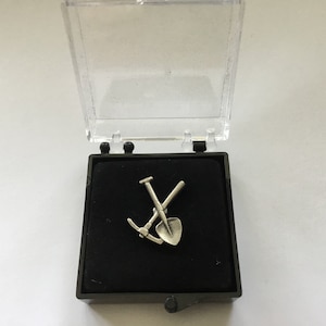 May include: A silver-colored pin in a black velvet box. The pin features a crossed pickaxe and shovel.