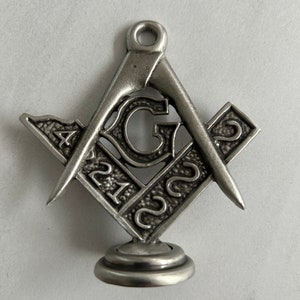Puede incluir: Un colgante de plata de escuadra y compás masónicos con una letra 'G' en el centro. El colgante tiene una superficie texturizada y un pequeño bucle en la parte superior para colgar.