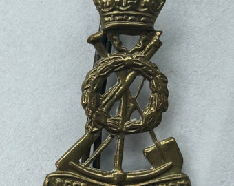 Puede incluir: Una insignia de metal dorado con una corona en la parte superior, una corona de laurel con rifles cruzados, un rifle, una pala y una pancarta con el texto "Labor Omnia Vincit".