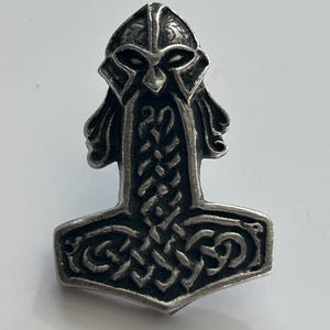 Może przedstawiać: Srebrny wisiorek w kształcie młota Thora, Mjolnira, z motywem hełmu wikinga. Wisiorek posiada skomplikowane sploty i szczegółową twarz. Młot ma około 3,8 cm wysokości.