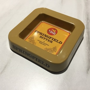 Op de afbeelding: Een vierkante, bruinbeige asbak met afgeronde randen. Het midden heeft een geel en oranje label met de tekst "SPRINGFIELD BITTER" en het "M&B" logo. De asbak is aan de zijkant gemarkeerd met "SPRINGFIELD".
