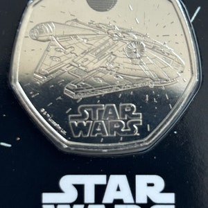 Puede incluir: Una moneda de color plateado con una imagen del Halcón Milenario, nave espacial de Star Wars. La moneda tiene la inscripción "Star Wars".