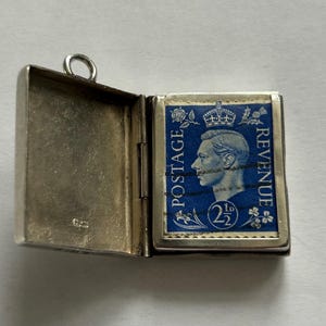 May include: Open silver locket with a blue postage stamp inside. The stamp features a profile of a man, the text "POSTAGE REVENUE", and the value "2½". The locket is rectangular with a hinged opening and a small loop at the top.