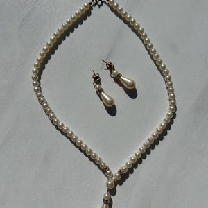 May include: A pearl necklace with a teardrop pendant and matching pearl earrings. The necklace is made of white pearls and has a gold clasp. The earrings are also made of white pearls and have gold posts.