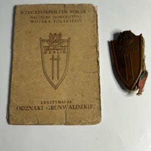 Puede incluir: Un documento y una insignia militares polacos antiguos. El documento es de color beige con texto polaco y un emblema de escudo. La insignia es de color bronce, en forma de escudo, y presenta texto y espadas cruzadas.
