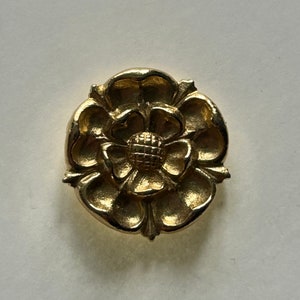 Puede incluir: Un pin de metal de color dorado en forma de rosa. La rosa tiene cinco pétalos y un centro con un diseño texturizado.