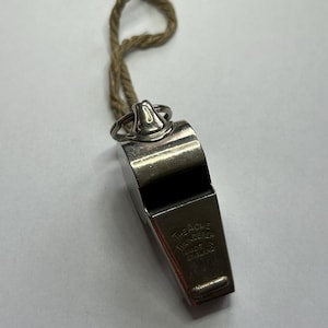 Puede incluir: Un silbato plateado con una boquilla negra y un cordón marrón. El silbato tiene el texto "The Acme Thunderer Made in England" grabado en el lateral.