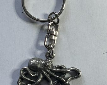 May include: Silver-colored keychain featuring an octopus charm. The octopus has detailed tentacles and a textured body. The keychain includes a split ring and a lobster clasp for easy attachment to keys or bags. The metal has a shiny finish.