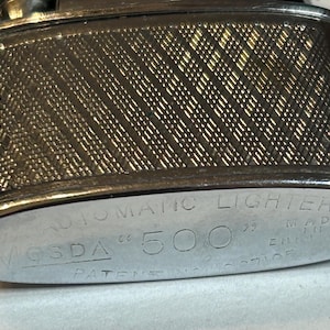 May include: Close-up of a vintage Vosda 500 automatic lighter. The lighter has a textured, diamond-patterned surface and a silver-colored metal finish. The text "AUTOMATIC LIGHTER", "VOSDA 500", and "MADE IN ENGLAND" are engraved on the lighter.