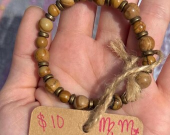 Puede incluir: Un brazalete de cuentas marrones y doradas con una cuerda marrón atada alrededor. El brazalete está en una mano y hay una etiqueta de precio que dice "10 €" y "Mm" en ella.