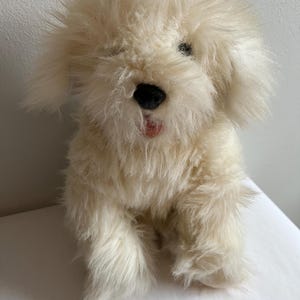 Op de afbeelding: Een crème-kleurig pluchen hondenspeelgoed met een pluizige vacht. Het speelgoed heeft zwarte ogen, een zwarte neus en een kleine roze tong. De hond zit rechtop.