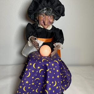 May include: A witch doll with gray hair, a black hat, and a black dress with an orange sash. The doll has green eyes, a large nose, and black fingernails. It sits behind a purple skirt featuring a yellow moon and star pattern, holding a glowing orange orb.