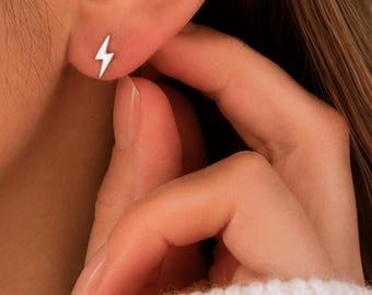 Puede incluir: Un pendiente de botón plateado con forma de rayo se muestra en una oreja. El pendiente es pequeño y sencillo, con un diseño minimalista. El pendiente es de color plateado brillante. El fondo está borroso, con el foco en el pendiente.