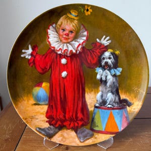 Könnte beinhalten: Dekorative Platte mit einem gemalten Bild eines Clowns in einem roten Kostüm mit weißem Rüschenkragen und einem kleinen Hund auf einem Zirkuspodest. Die Platte hat einen goldenen Rand und einen bemalten Hintergrund.