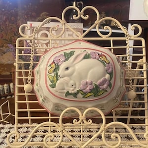 May include: Cream-coloured metal cookbook stand with a decorative, oval ceramic plaque. The plaque depicts a white rabbit and baby rabbit, surrounded by pink flowers and butterflies. The stand has a heart-shaped design.