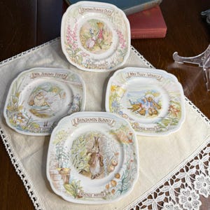 May include: Four square-shaped decorative plates featuring illustrations from Beatrix Potter stories. Each plate has a different character: Jemima Puddle-Duck, Mr. Jeremy Fisher, Mrs. Tiggy-Winkle, and Benjamin Bunny. The plates are white with colorful images and floral borders.
