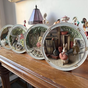 Puede incluir: Una colección de platos decorativos con escenas de conejos en un jardín. Los platos tienen un diseño en relieve con colores detallados y un borde blanco. Los platos se exhiben sobre una superficie de madera.