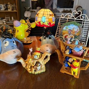 Könnte beinhalten: Eine Sammlung von Teekannen und Figuren zum Thema Winnie Puuh. Die Teekannen sind wie Eeyore, Tigger und ein Baumstumpf geformt. Die Figuren umfassen Winnie Puuh, Ferkel und andere Charaktere. Eine Buntglaslampe ist ebenfalls zu sehen.