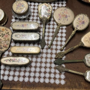 Pode incluir: Uma coleção de acessórios de toucador vintage, incluindo vários espelhos de mão, escovas de cabelo e um pequeno frasco com tampa. Cada peça apresenta uma moldura de metal dourado ornamentada e um encaixe creme com bordados florais.