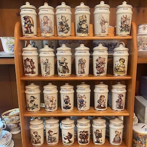Può includere: Una rastrelliera in legno con 20 barattoli di spezie in ceramica. Ogni barattolo è etichettato con il nome di una spezia e presenta un'illustrazione stravagante. Le spezie includono coriandolo, timo e zenzero.