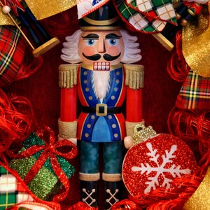 Puede incluir: Un soldado cascanueces de madera con uniforme rojo, azul y dorado, con sombrero negro y pelo blanco. Un adorno de copo de nieve rojo brillante y una caja de regalo verde brillante est&aacute;n en primer plano, rodeados de elementos decorativos rojos y dorados.