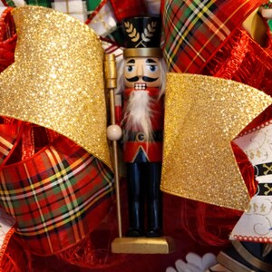 Puede incluir: Una corona navide&ntilde;a festiva con un soldado cascanueces de madera con barba blanca, uniforme rojo y negro y detalles dorados. La corona incluye lazos dorados con purpurina y cintas rojas a cuadros.