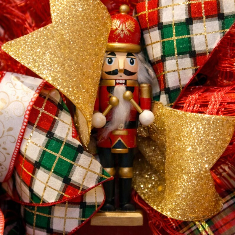 Puede incluir: Decoraci&oacute;n navide&ntilde;a festiva con un soldado cascanueces de madera con uniforme rojo y negro, barba y bigote blancos. El cascanueces est&aacute; rodeado de lazos dorados con purpurina y cintas a cuadros rojas, verdes y blancas.