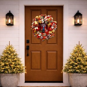 Puede incluir: Una festiva corona navide&ntilde;a con un cascanueces en el centro cuelga de una puerta marr&oacute;n. La corona presenta cintas rojas, doradas y verdes, y est&aacute; flanqueada por dos peque&ntilde;os &aacute;rboles de Navidad en macetas, iluminados con luces blancas c&aacute;lidas.