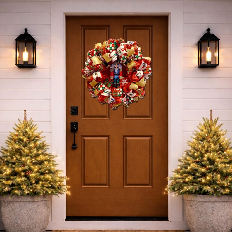 Puede incluir: Una festiva corona navide&ntilde;a con un cascanueces en el centro cuelga de una puerta marr&oacute;n. La corona presenta cintas rojas, doradas y verdes, y est&aacute; flanqueada por dos peque&ntilde;os &aacute;rboles de Navidad en macetas, iluminados con luces blancas c&aacute;lidas.