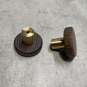 Può includere: Due pomelli per mobili. Uno assemblato, con una faccia rotonda in legno scuro e uno stelo in ottone. L'altro è smontato, con un cilindro in ottone su un disco di legno scuro. I pomelli sono su una superficie grigia.