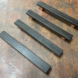 May include: Four dark gray wooden cabinet pulls with copper-colored end caps are arranged diagonally on a brown surface. The pulls have a rectangular shape and a wood grain texture, suitable for kitchen or bathroom cabinets.